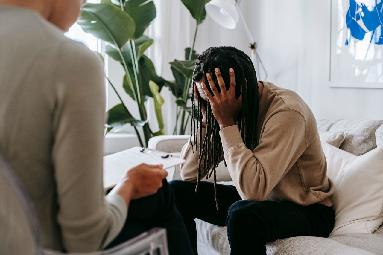 Anxious African American male with hands on head sitting on sofa near crop female psychologist with clipboard during appointment in office